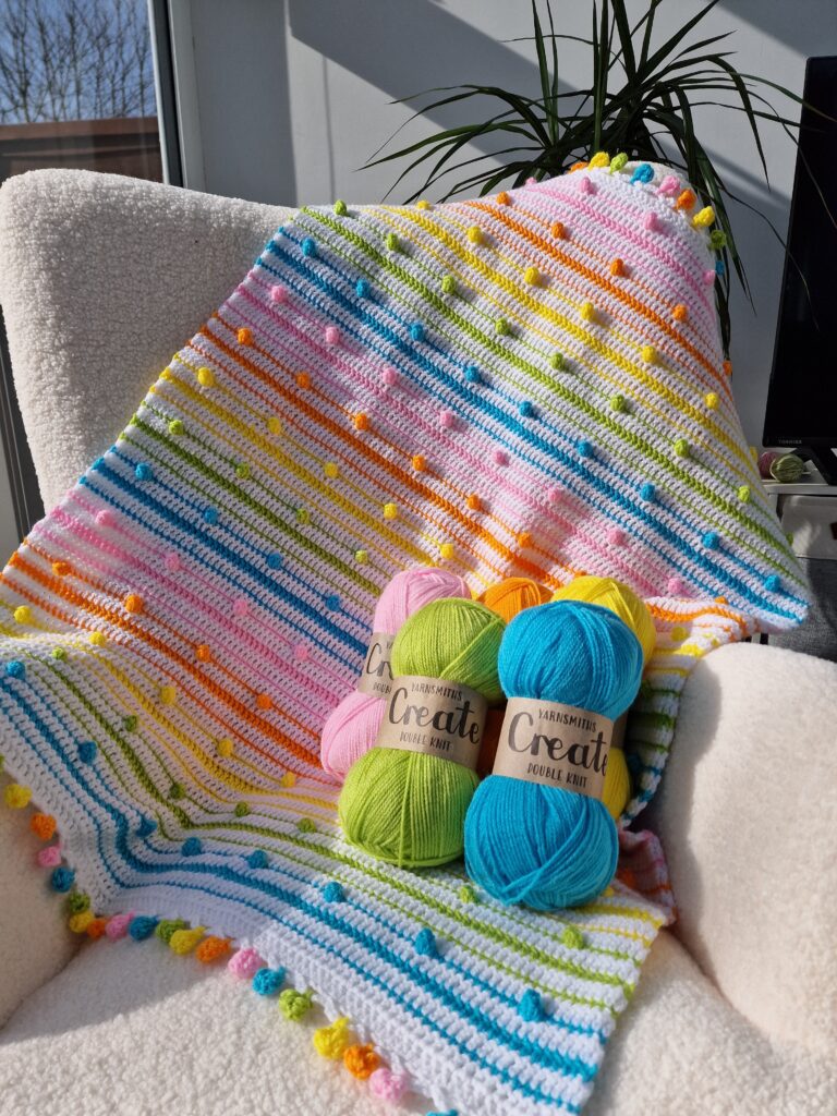 Close-up of the 'A Spot of Spring' crochet blanket by EmkatCrochet, featuring a detailed floral-inspired pattern in soft pastel yarns, draped gently to show texture and stitch work. Yarn is yarnsmiths create dk from wool warehouse.