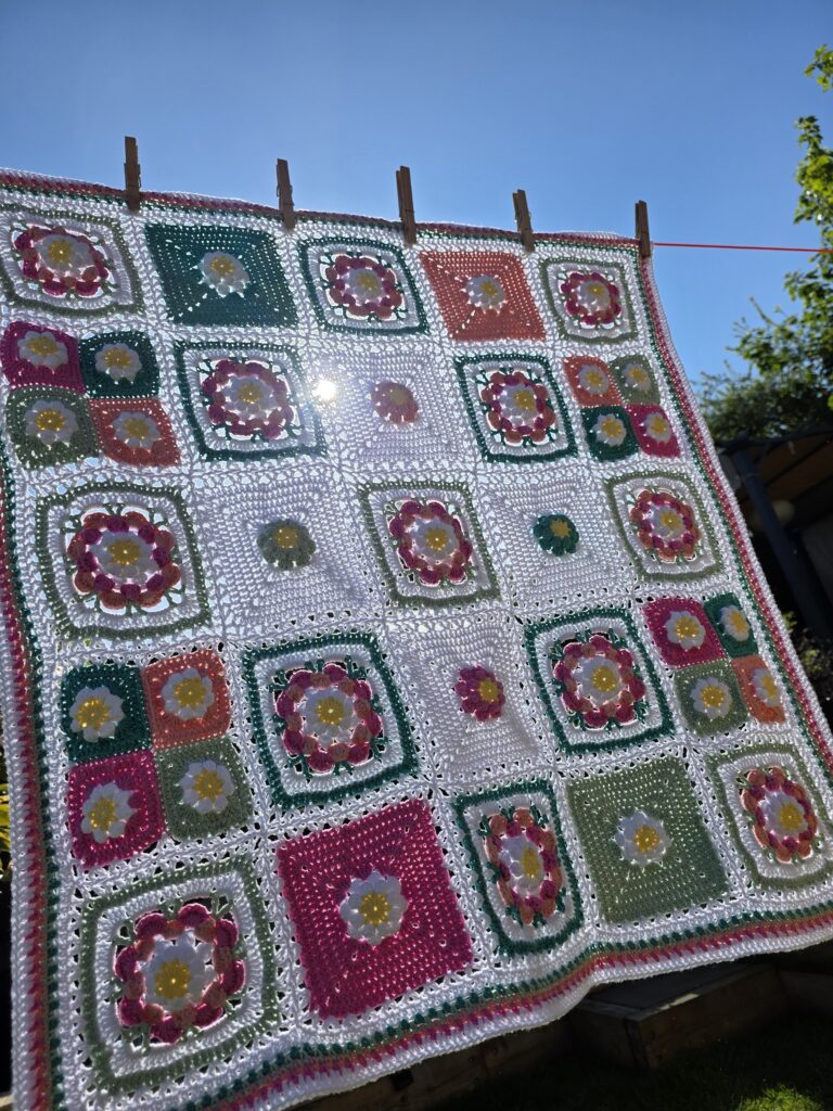 A colorful crocheted patchwork blanket with floral granny squares draped over a wicker chair, with skeins of yarn in pink, yellow, green, teal, and peach resting on it, set outdoors in front hanging off the washing line.