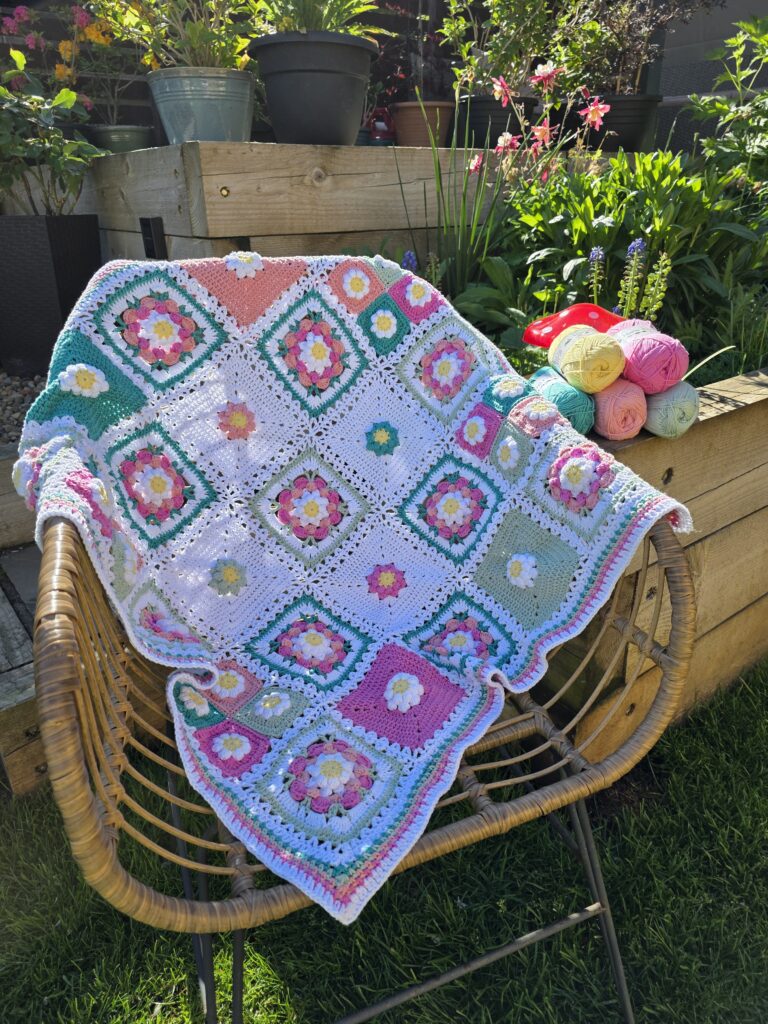 A colorful crocheted patchwork blanket with floral granny squares draped over a wicker chair, with skeins of yarn in pink, yellow, green, teal, and peach resting on it, set outdoors in front of a wooden shed.