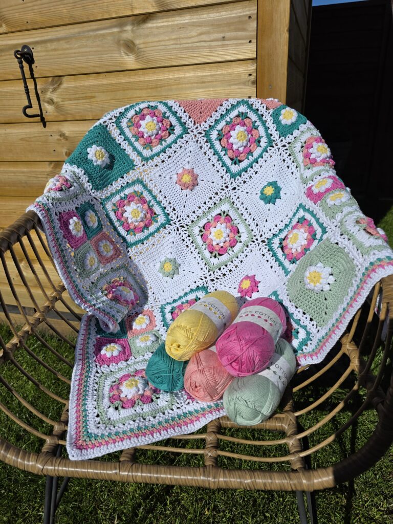 A colorful crocheted patchwork blanket with floral granny squares draped over a wicker chair, with skeins of yarn in pink, yellow, green, teal, and peach resting on it, set outdoors in front of a wooden shed.