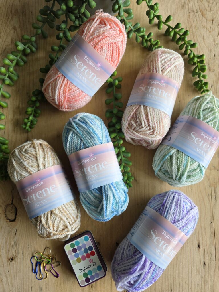 Small 25g balls of stylecraft serene aran yarn, each made with soft, pastel-shaded yarns in colors like pink, blue, green, tan, and purple. The yarn used appears to be Stylecraft New Serene Aran, known for its variegated, softly blended tones. Small matching yarn balls sit in the center, and trailing succulent plants and a crocheted stone cover add a natural decorative touch