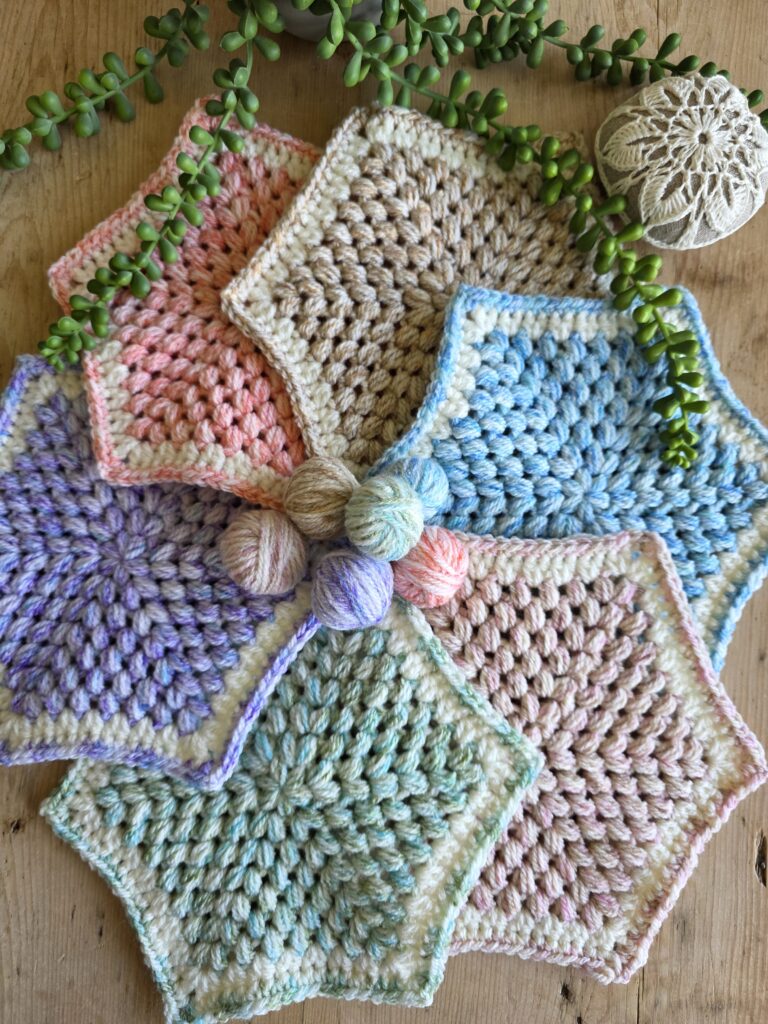 Hexagonal crochet swatches arranged in a circular layout, each made with soft, pastel-shaded yarns in colors like pink, blue, green, tan, and purple. The yarn used appears to be Stylecraft New Serene Aran, known for its variegated, softly blended tones. Small matching yarn balls sit in the center, and trailing succulent plants and a crocheted stone cover add a natural decorative touch