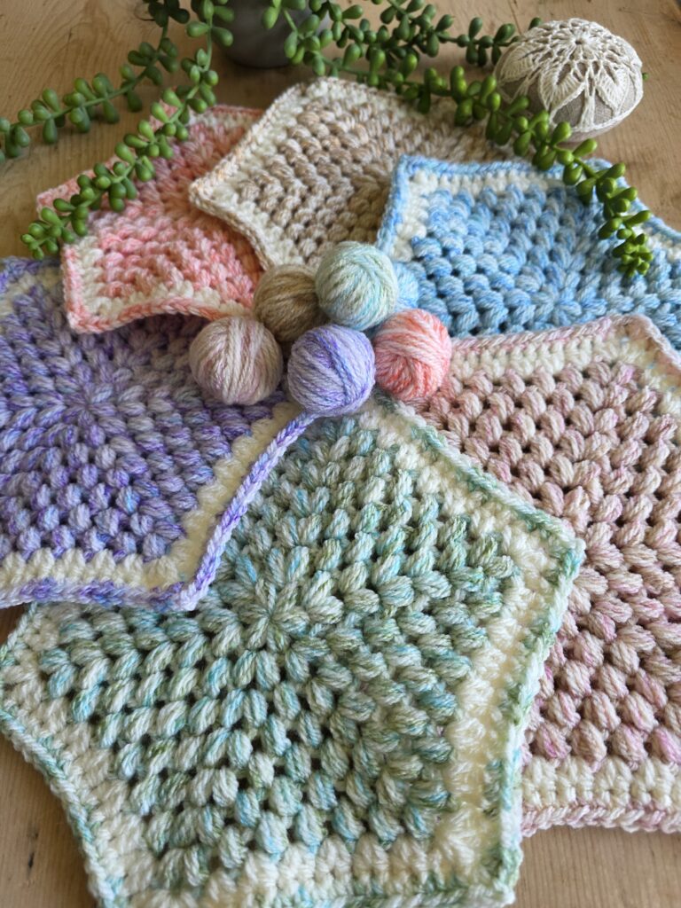 Hexagonal crochet swatches arranged in a circular layout, each made with soft, pastel-shaded yarns in colors like pink, blue, green, tan, and purple. The yarn used appears to be Stylecraft New Serene Aran, known for its variegated, softly blended tones. Small matching yarn balls sit in the center, and trailing succulent plants and a crocheted stone cover add a natural decorative touch