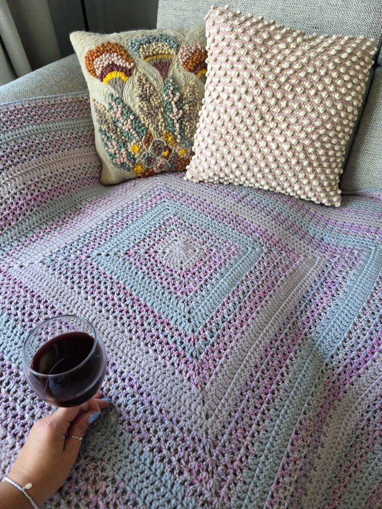 A cosy crochet setup featuring a square blanket worked in the round with soft pastel stripes of grey, pink, and blue tones, draped over a light grey couch. Two textured pillows rest against the back of the couch—one with a raised bobble stitch in pale pink and cream, and another with a colorful floral embroidery design. A hand holding a glass of red wine is visible in the bottom left corner, suggesting a relaxed, comforting atmosphere.