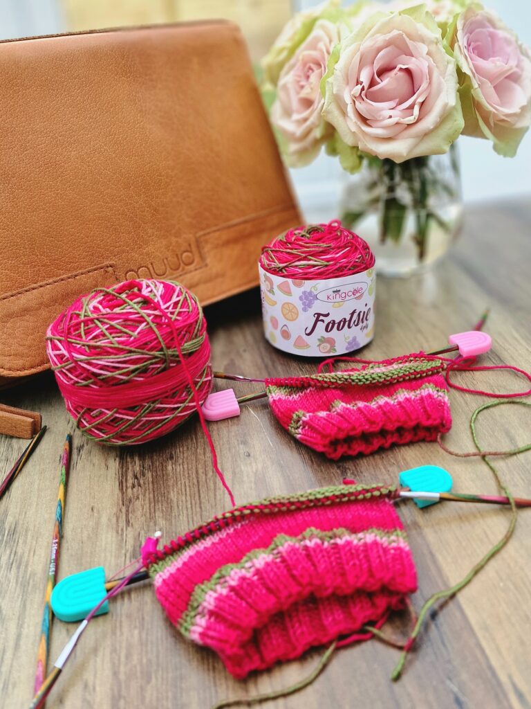 Hand-knitted socks in King Cole Footsie yarn, Watermelon colour, on KnitPro circular needles with a Muud Living tan leather project bag
