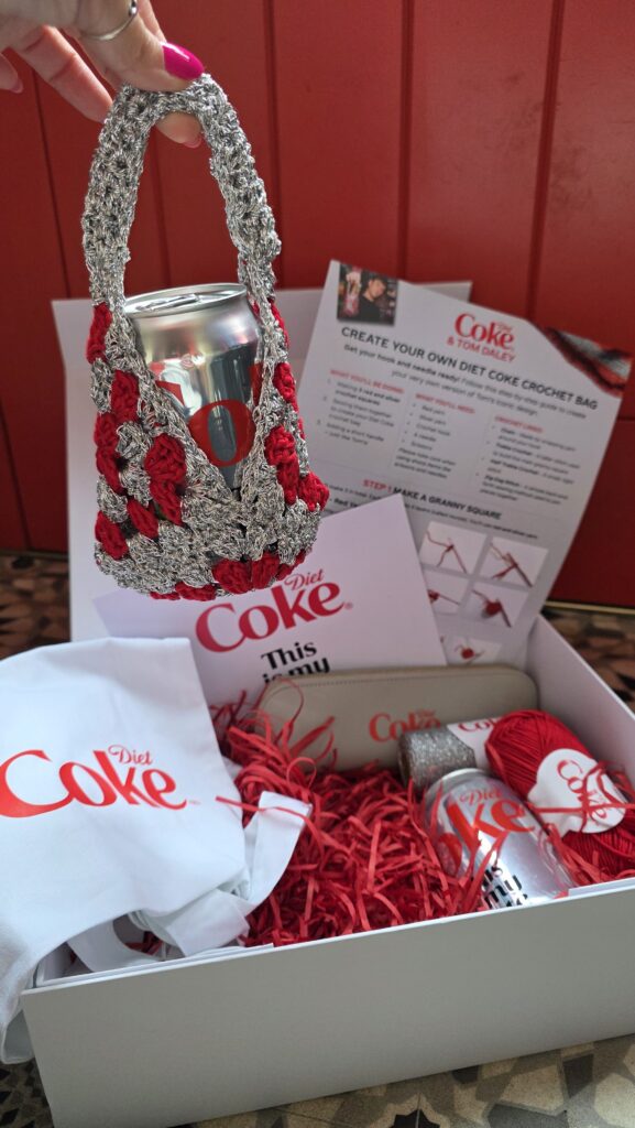 Diet Coke gift box containing two cans, a crochet bag, crochet hook, tote bag, and skeins of red and silver yarn arranged neatly in a presentation box
