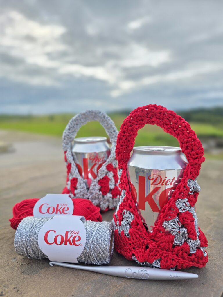 Two Diet Coke cans in crochet bags with a Diet Coke-themed crochet hook hanging on a wall, fields visible in the background