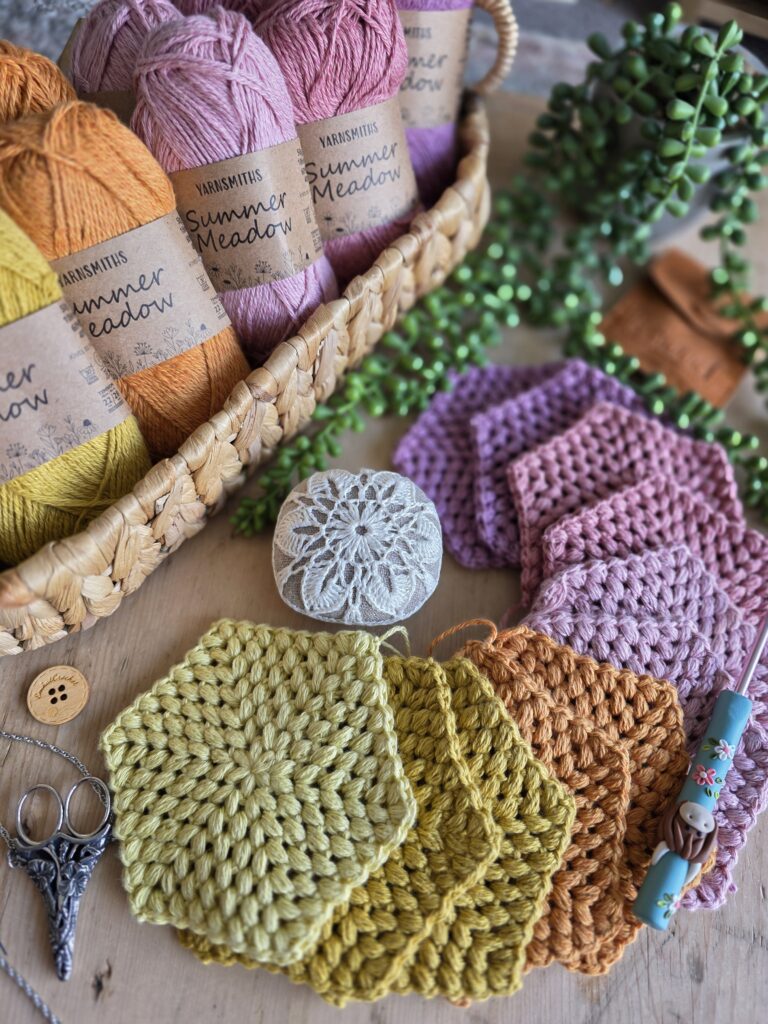 pink, orange and yellow Summer Meadow puff stitch hexagons displayed with Yarnsmiths DK yarn, a crochet pincushion, and polymer clay crochet hook by PedrosPlaques in a wicker basket