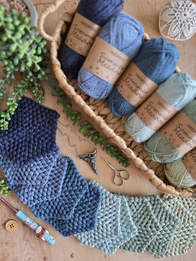 Handmade puff stitch hexagons in blue and green from the Yarnsmiths Summer Meadow DK range, with yarn balls in a wicker basket, crochet pincushion, and PedrosPlaques polymer clay hook