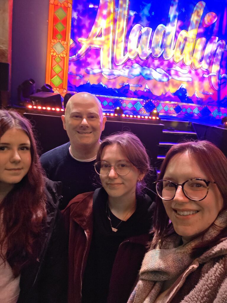 Family selfie taken at the theatre during a performance of Aladdin, smiling under colourful stage lights.