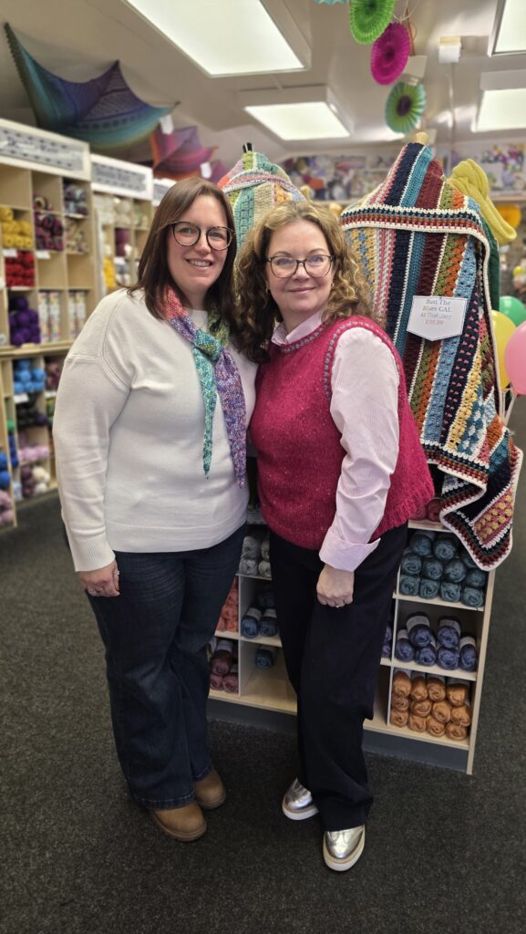 Two women standing together inside a yarn shop, surrounded by shelves of colourful yarn and crochet samples.