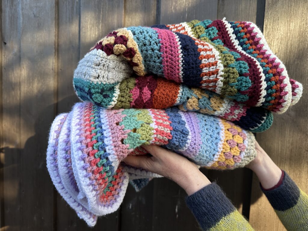 Stack of colourful crochet blankets folded and held up to show textured stitch patterns and bright stripes.